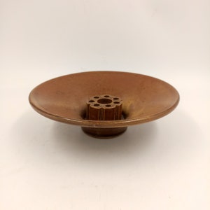 May include: A brown ceramic bowl with a small, round, brown ceramic base. The bowl has a wide, shallow opening and a central hole with multiple smaller holes around it.
