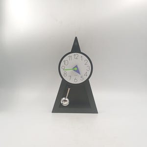 Puede incluir: Un reloj de escritorio triangular negro con una esfera blanca con números negros y detalles en verde y azul. Un péndulo plateado cuelga debajo. El reloj tiene un diseño moderno y minimalista.