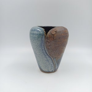 May include: A ceramic vase with a unique heart shape, featuring a blue and brown glaze. The vase has a textured surface and a slightly uneven opening.