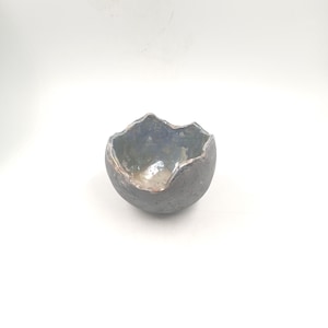 Peut inclure: Un petit bol en céramique fait à la main, avec un extérieur gris foncé et un intérieur bleu clair. Le bol a un bord irrégulier et texturé et une forme arrondie. L'intérieur a une finition brillante, sur fond blanc.