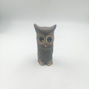 Peut inclure: Petite figurine de hibou marron avec de grands yeux sombres et une surface texturée. Le hibou a une posture simple et droite et de petits pieds beiges. La figurine est probablement en céramique ou dans un matériau similaire, sur fond blanc.
