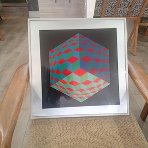 May include: Framed geometric art print featuring a 3D cube design. The cube is composed of red, green, and blue diamond shapes against a black background. The artwork is encased in a white frame and rests on a chair.