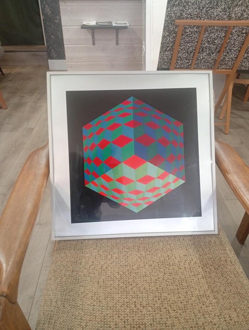 May include: Framed geometric art print featuring a 3D cube design. The cube is composed of red, green, and blue diamond shapes against a black background. The artwork is encased in a white frame and rests on a chair.