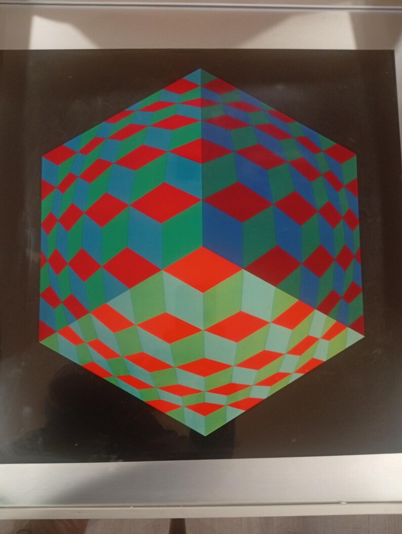 May include: A framed geometric art print featuring a three-dimensional cube design. The cube is composed of red, green, and blue diamond shapes, creating an optical illusion against a black background. The artwork is framed in a silver metal frame.
