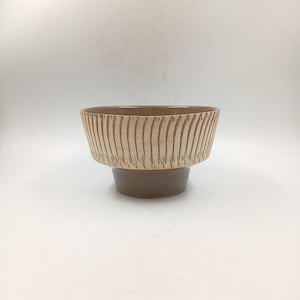 Peut inclure: Un bol en céramique marron avec un bord texturé de couleur claire orné de lignes verticales. Le bol repose sur une base plus courte, marron foncé. Le design est minimaliste et moderne, adapté à un usage décoratif ou fonctionnel.