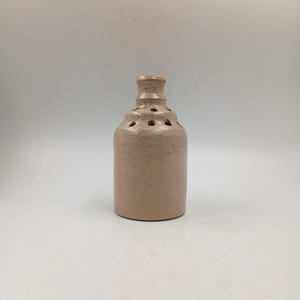 Peut inclure: Une bouteille en céramique marron clair mouchetée, avec un col étroit et une série de trous circulaires sur le corps. La bouteille a une forme cylindrique et repose sur un fond blanc. La céramique a une texture rugueuse.