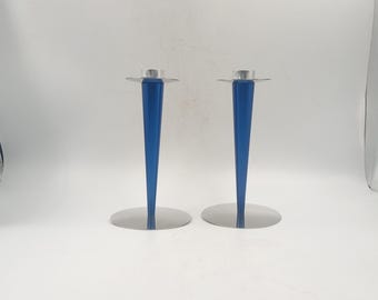 May include: Two blue and silver candle holders. Each holder has a conical blue stem and a circular silver base and top. The candle holders are displayed on a white background.