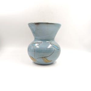 Op de afbeelding: Een lichtblauwe keramische vaas met een bolvormig lichaam en een uitlopende rand. De vaas heeft een handgeschilderd bloemmotief met lichtblauwe en gouden accenten. De vaas is ongeveer 20 cm hoog en zou een decoratieve toevoeging zijn aan elke kamer.