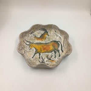 May include: A decorative bowl with a scalloped edge, featuring a beige and brown speckled finish. The bowl showcases a hand-painted scene of prehistoric animals in yellow, orange, and black. The design of the bowl evokes a cave painting aesthetic.