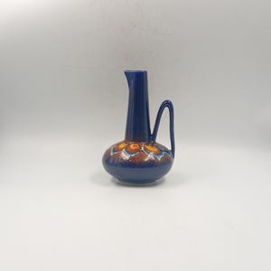 Peut inclure: Pichet en céramique bleu foncé avec une base bulbeuse et un col long et fin. La base présente un motif floral dans les tons orange, marron et bleu. Il possède une anse incurvée et un bec verseur.