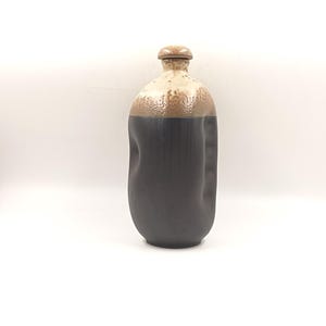 Puede incluir: Una botella de cerámica negra y blanca con una tapa marrón. La botella tiene una forma única e irregular.