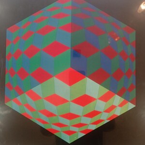 May include: Geometric artwork featuring a three-dimensional cube design. The cube is composed of red, green, and blue diamond shapes, creating an optical illusion against a black background. The artwork is framed.