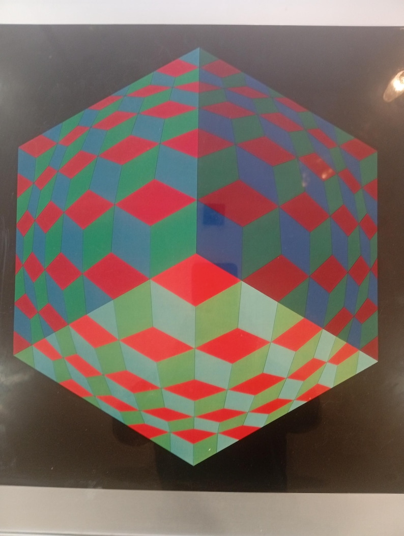 May include: Geometric artwork featuring a three-dimensional cube design. The cube is composed of red, green, and blue diamond shapes, creating an optical illusion against a black background. The artwork is framed.
