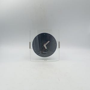 May include: A contemporary clock featuring a black clock face with silver hands, set within a clear acrylic casing. The design includes silver metal detailing. The word "Tetra" is printed at the base of the clock face.
