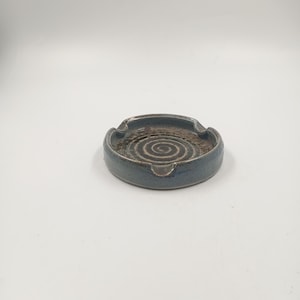 Puede incluir: Un cenicero de cerámica con un esmalte azul oscuro. El cenicero tiene forma circular con un centro empotrado con un diseño en espiral. Tiene tres pequeñas muescas para apoyar los cigarrillos.