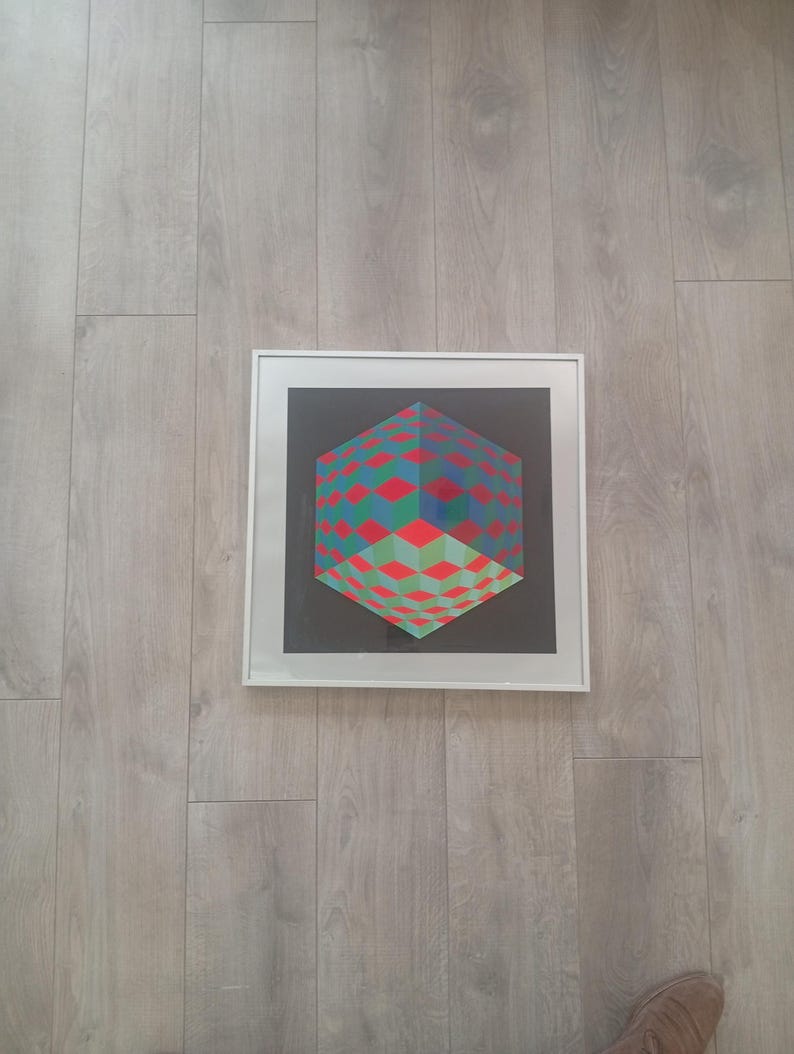May include: Framed geometric art print featuring a 3D cube design. The cube is composed of red, green, and blue diamond shapes against a black background. The artwork is encased in a white frame.