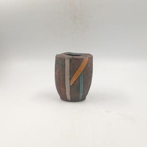 Peut inclure: Petit vase en céramique marron avec un motif géométrique. Le vase a un dessus carré et un corps cylindrique. Des rayures verticales orange, bleues et blanches ornent l'extérieur. Le vase est probablement fait à la main et convient aux petits arrangements floraux ou à un usage décoratif.
