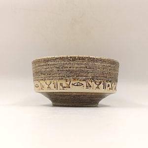 Può includere: Una piccola ciotola in ceramica marrone con una fascia bianca attorno al bordo superiore. La fascia è decorata con un motivo ripetitivo di simboli stilizzati.