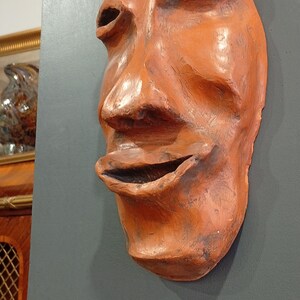 Pode incluir: Uma m&aacute;scara de terracota com uma boca larga e aberta e tra&ccedil;os exagerados. A m&aacute;scara &eacute; de cor laranja intensa e tem uma superf&iacute;cie texturizada.