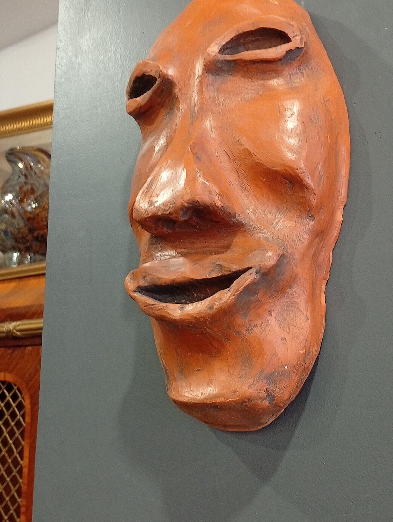 Pode incluir: Uma m&aacute;scara de terracota com uma boca larga e aberta e tra&ccedil;os exagerados. A m&aacute;scara &eacute; de cor laranja intensa e tem uma superf&iacute;cie texturizada.
