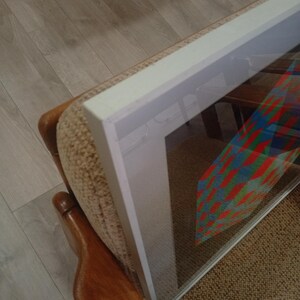 May include: Framed artwork with a colorful geometric design, displayed on a beige upholstered chair. The frame is white, and the artwork features red, blue, and green patterns. The chair has wooden armrests and is positioned on a light brown carpet.