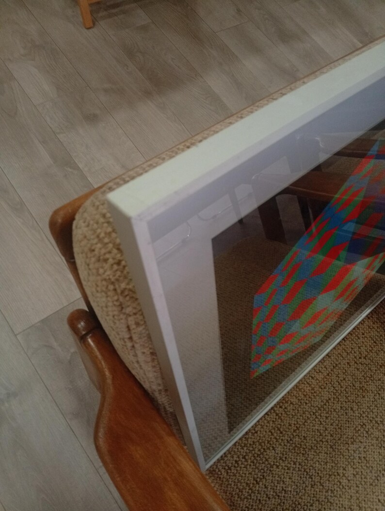 May include: Framed artwork with a colorful geometric design, displayed on a beige upholstered chair. The frame is white, and the artwork features red, blue, and green patterns. The chair has wooden armrests and is positioned on a light brown carpet.