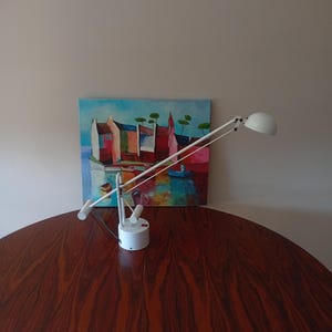 May include: A white desk lamp with a rounded shade and adjustable arm is positioned on a dark wood table. A colorful painting of buildings and boats hangs on the wall behind the lamp.