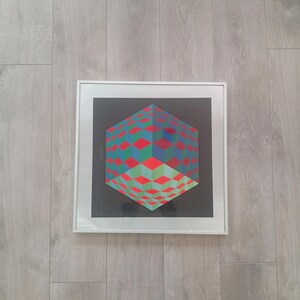 May include: A framed geometric art print featuring a three-dimensional cube design. The cube is composed of red, green, and blue diamond shapes against a black background. The artwork is encased in a white frame and mounted on a light wood-grain surface.