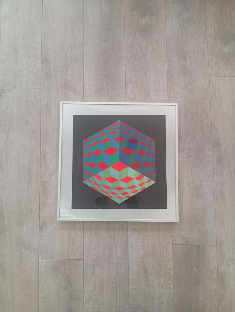 May include: A framed geometric art print featuring a three-dimensional cube design. The cube is composed of red, green, and blue diamond shapes against a black background. The artwork is encased in a white frame and mounted on a light wood-grain surface.