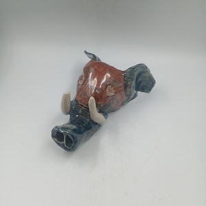 Peut inclure: Sculpture en céramique représentant une tête de sanglier. La tête est principalement brun-rouge avec des détails bleus et deux défenses ivoire. La sculpture a une surface texturée et détaillée, sur fond blanc.