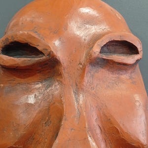 Pode incluir: Uma escultura de terracota de um rosto com tra&ccedil;os exagerados. O rosto tem olhos grandes e ocos e um nariz proeminente.