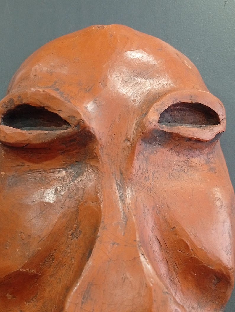 Pode incluir: Uma escultura de terracota de um rosto com tra&ccedil;os exagerados. O rosto tem olhos grandes e ocos e um nariz proeminente.