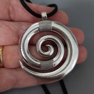 Op de afbeelding: Zilverkleurige spiraalvormige hangerketting met een zwart koord. De hanger heeft een groot, open spiraalontwerp met een getextureerde afwerking. De spiraal heeft een diameter van ongeveer 5 cm en het koord is een eenvoudig, rond zwart materiaal.