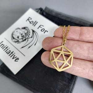 Pode incluir: Um colar de tom dourado com um pingente geométrico de estrutura aberta em forma de dado de vinte lados. O pingente está suspenso por uma corrente dourada fina. Ao lado, há um cartão branco com o texto preto Roll For Initiative e um desenho de dragão.