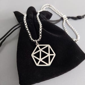 Può includere: Una collana d'argento con un ciondolo geometrico. Il ciondolo è un esagono con un design geometrico all'interno. La collana è su una pochette di velluto nero.