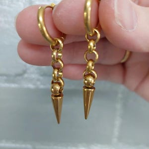 Könnte beinhalten: Ein Paar goldfarbene Creolen mit hängendem Kettendesign. Jeder Ohrring besteht aus einem runden Reifen, der mit einer kurzen Kette aus drei kleinen Ringen verbunden ist und in einer glatten Kugel sowie einem spitzen, kegelförmigen Anhänger endet.