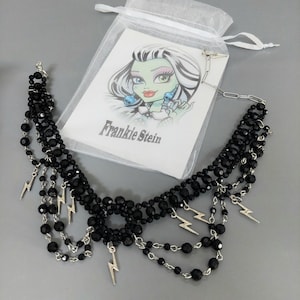 Op de afbeelding: Een meerlaagse ketting met zwarte gefacetteerde kralen en zilverkleurige bliksemschichtbedels aan een zilveren ketting. De ketting wordt getoond met een witte kaart met een illustratie van Frankie Stein en een wit organza zakje.