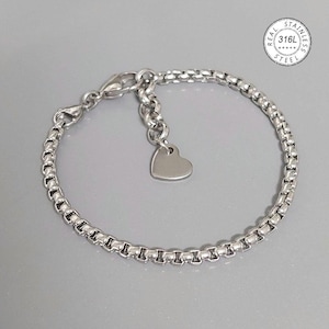 Op de afbeelding: Een zilverkleurige armband met een box chain design en een hartvormige bedel. De armband heeft een karabijnsluiting en een verstelbare ketting. De afbeelding bevat een ronde stempel met de tekst "REAL STAINLESS STEEL 316L".