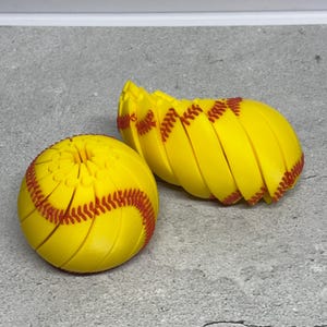 Puede incluir: Dos pelotas de sóftbol amarillas con costuras rojas. Una está entera y la otra está separada en secciones. Las pelotas de sóftbol están sobre una superficie gris. Las pelotas de sóftbol están diseñadas para parecerse a pelotas de sóftbol reales.