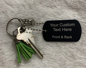 Könnte beinhalten: Ein schwarzer, rechteckiger Schlüsselanhänger mit dem Text "Your Custom Text Here Front & Back" ist an einem Schlüsselring mit Schlüsseln und einer grünen Quaste befestigt. Das Etikett ist mit einer kleinen Kugelkette mit dem Ring verbunden.