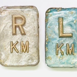 May include: Two small, rectangular resin tiles with gold lettering. The tile on the left is white with gold glitter and says "R KM". The tile on the right is blue with gold glitter and says "L KM".