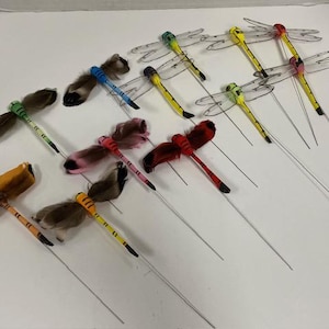 May include: A collection of ten dragonfly-shaped ornaments featuring long, thin bodies in various colors including yellow, red, blue, and orange. Each dragonfly has wings made of either translucent plastic or soft, textured feathers attached to a wire stem.