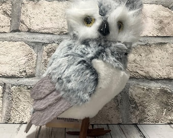 Puede incluir: Un búho de peluche gris y blanco con patas marrones. El búho tiene ojos grandes y redondos con pupilas amarillas. El búho está sentado sobre una superficie de madera.