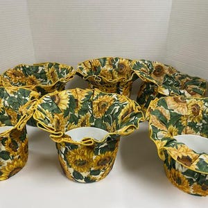 May include: A set of six fabric bowl cozies featuring a sunflower pattern with yellow petals and green leaves on a white background. Each cozy is constructed with a ruffled edge and holds a white ceramic bowl, designed for holding hot items.