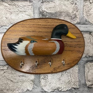 May include: Wooden key holder shaped like a duck with a painted design. The duck has a black head, brown body, and white and gray feathers. The key holder has four hooks.