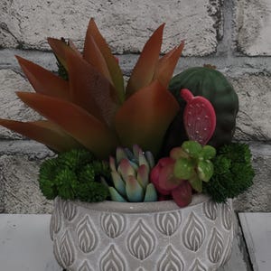 May include: A decorative succulent arrangement in a gray, patterned pot. The arrangement features various faux succulents in shades of green, red, and blue. The pot is round with a textured surface and sits on a white wooden surface.