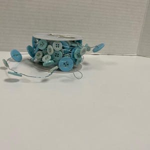 May include: A spool of light blue and white buttons on a string. The buttons are various sizes and shapes. Some buttons are round, and some are rectangular. The buttons are on a white surface.