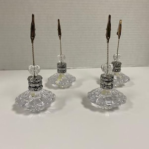 May include: Four clear glass photo holders with silver accents. Each holder features a decorative base, a beaded stem, and a metal clip at the top. The bases have a skirt-like design. The holders are arranged on a white surface.