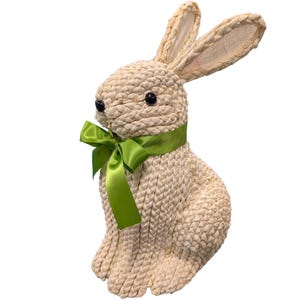 May include: A woven rabbit figurine with a green satin ribbon around its neck. The rabbit has black eyes and long, upright ears. The figurine is made of a light beige material, and is likely a decorative Easter or spring-themed item.