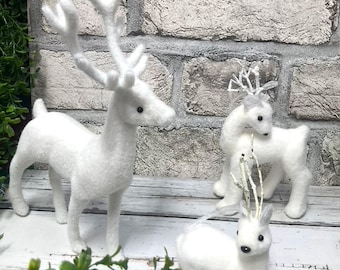 Puede incluir: Tres figuritas de reno blancas y peludas, una de pie con cuernos grandes, otra de pie con cuernos más pequeños y otra acostada. Los renos están sobre una superficie de madera blanca con follaje verde en primer plano.